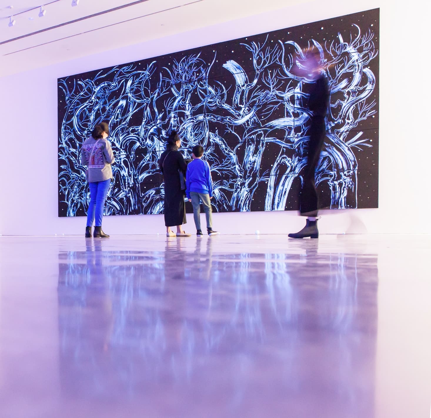 4 people standing in front of a graffiti painting. 1 woman looking down on her phone. A mother and son looking at the painting. A person blurred and in motion